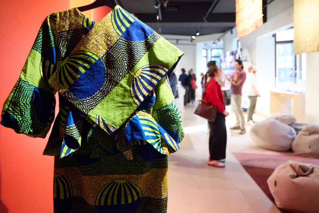 A blue and green dress hangs from the ceiling of a museum room. People visiting the museum can be seen on the background. The photo ilustrates the discussion about fashion.